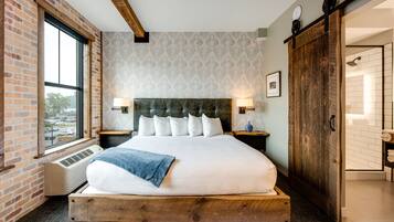 A large bed with a wooden frame, a headboard with multiple pillows, bedside lamps, a window with a view of the outside, and a brick wall.