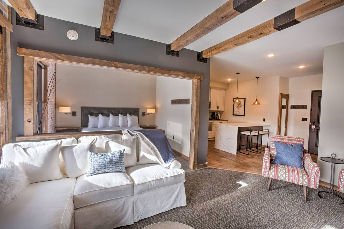 Quarto de hotel moderno com uma cama grande, um sofá branco, uma poltrona estampada e uma área de cozinha visível através de uma porta aberta.