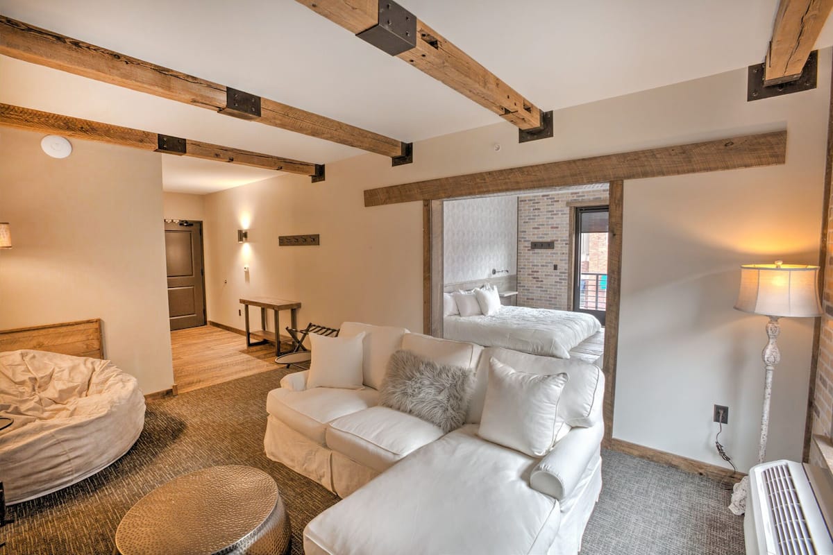 Quarto de hotel com sofá branco, uma cadeira redonda de vime, cama e vigas de madeira expostas no teto.