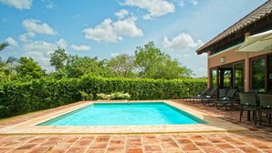 A rectangular swimming pool with a tiled deck, surrounded by lush greenery and a clear blue sky.
