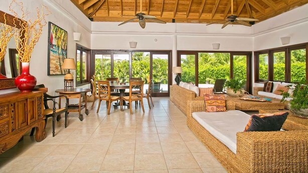 A spacious room with a high ceiling, wooden beams, a dining area with a table and chairs, a sofa set, and large windows offering a view of greenery.