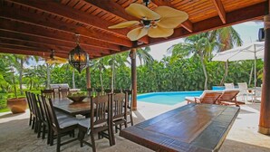 Un área de comedor al aire libre con mesas y sillas de madera, una piscina y palmeras al fondo.