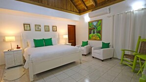 Un dormitorio con cama, dos sillas, una mesita de noche, lámpara, cuadro y ventana con cortinas.