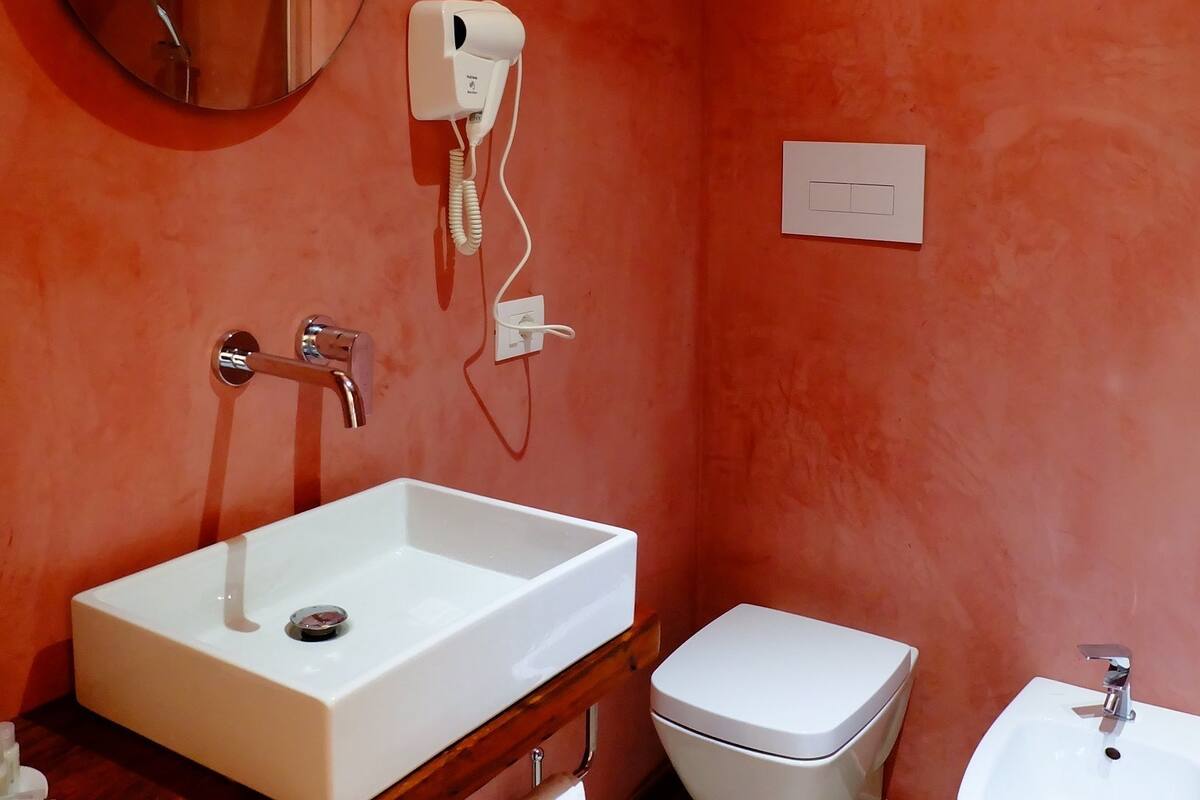 A bathroom with a white sink, a round mirror, a handheld hair dryer, a toilet, and a bidet.