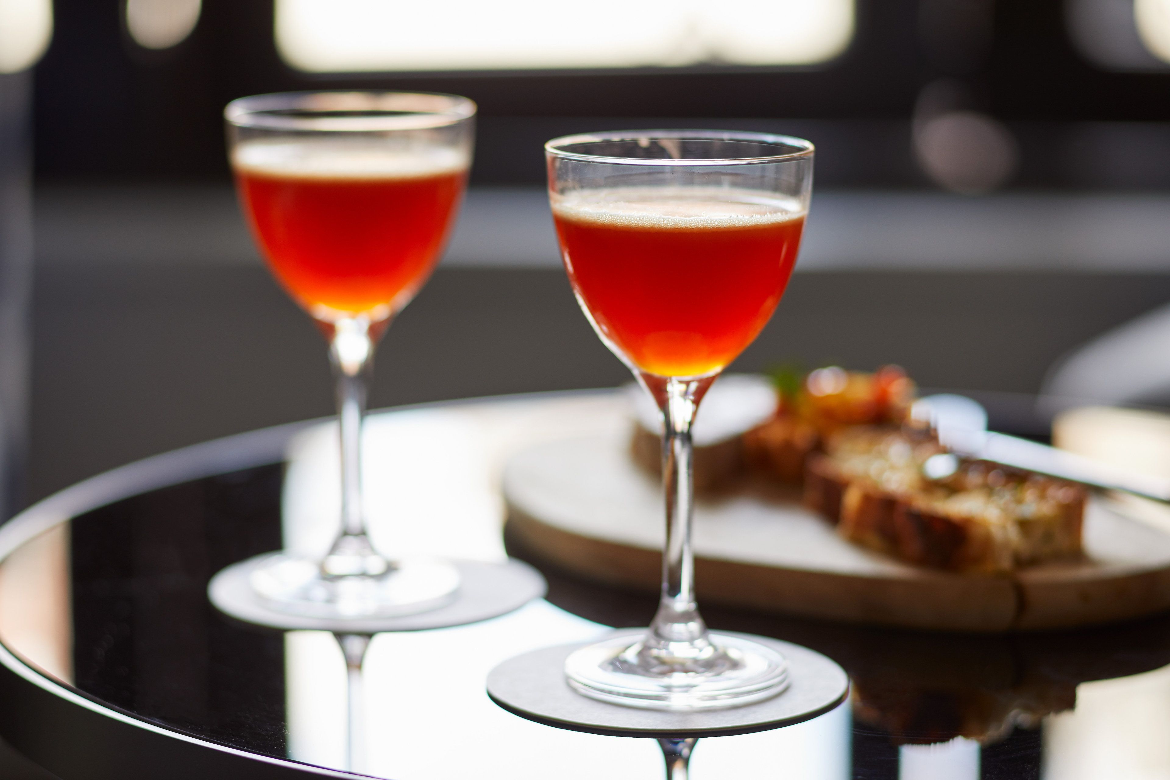 Two glasses of red beverage on a reflective surface with a plate of toast in the background.