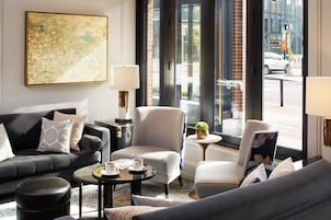 A modern hotel room with a sofa, armchairs, a coffee table, and a large window offering a city view.