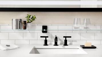 A modern bathroom with a white sink, black faucets, a glass shelf with toiletries, and a potted plant.