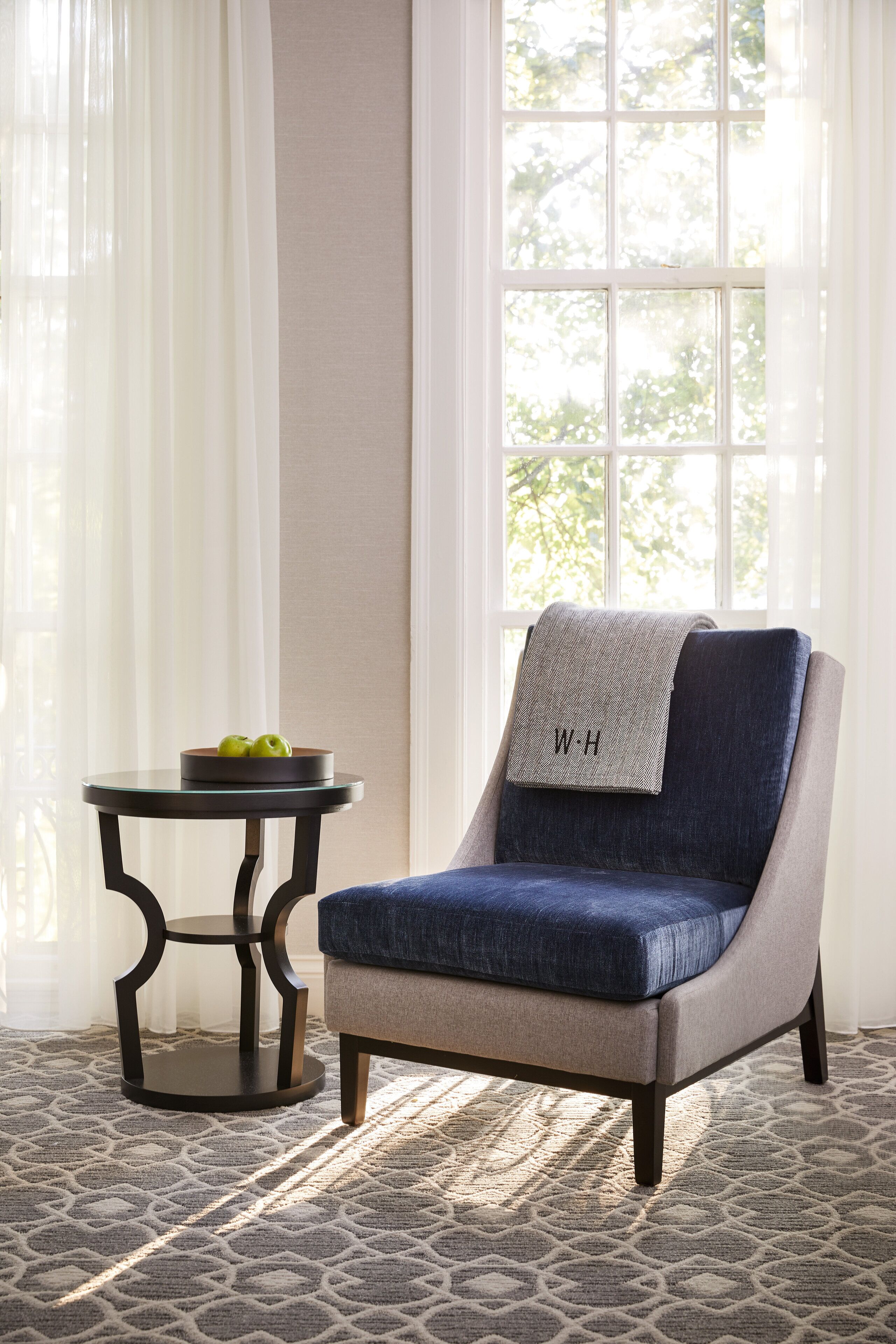A room with a blue armchair, a small round table with a bowl of apples, and a patterned rug.
