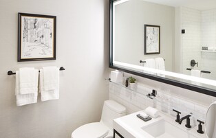 Ein modernes Badezimmer mit einem großen Spiegel, weißen Fliesen, einer Toilette, einem Waschbecken mit Glasablage und zwei gerahmten Kunstwerken an der Wand.