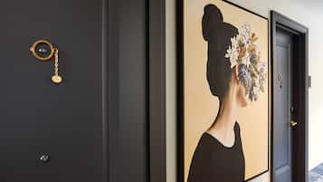 Een gang met een donkere deur, een lijstwerk met een afbeelding van een vrouw met bloemen, en een rond object aan de muur.