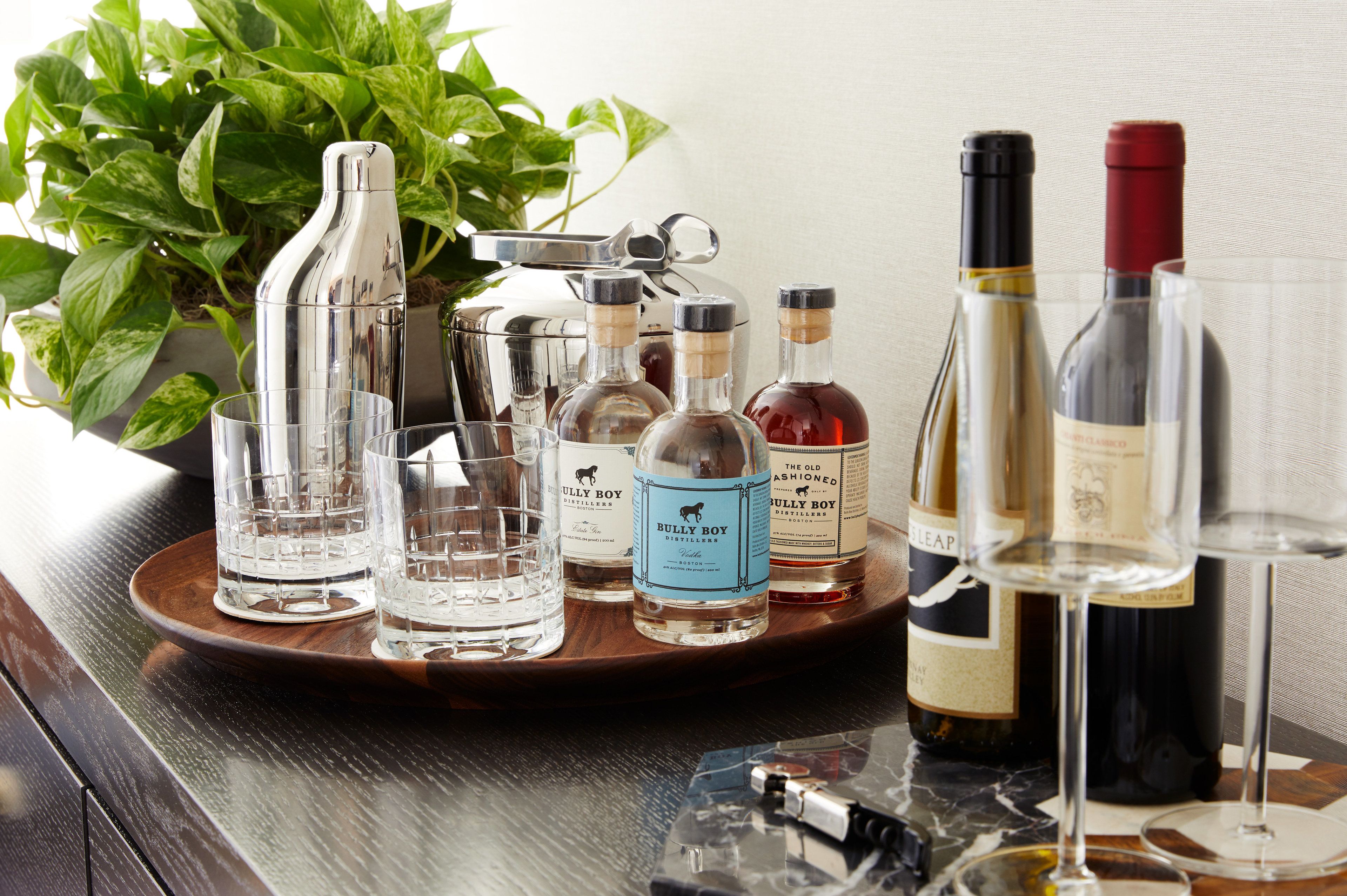 A tray with various bottles and glasses, likely for serving drinks, placed on a wooden table with a potted plant in the background.