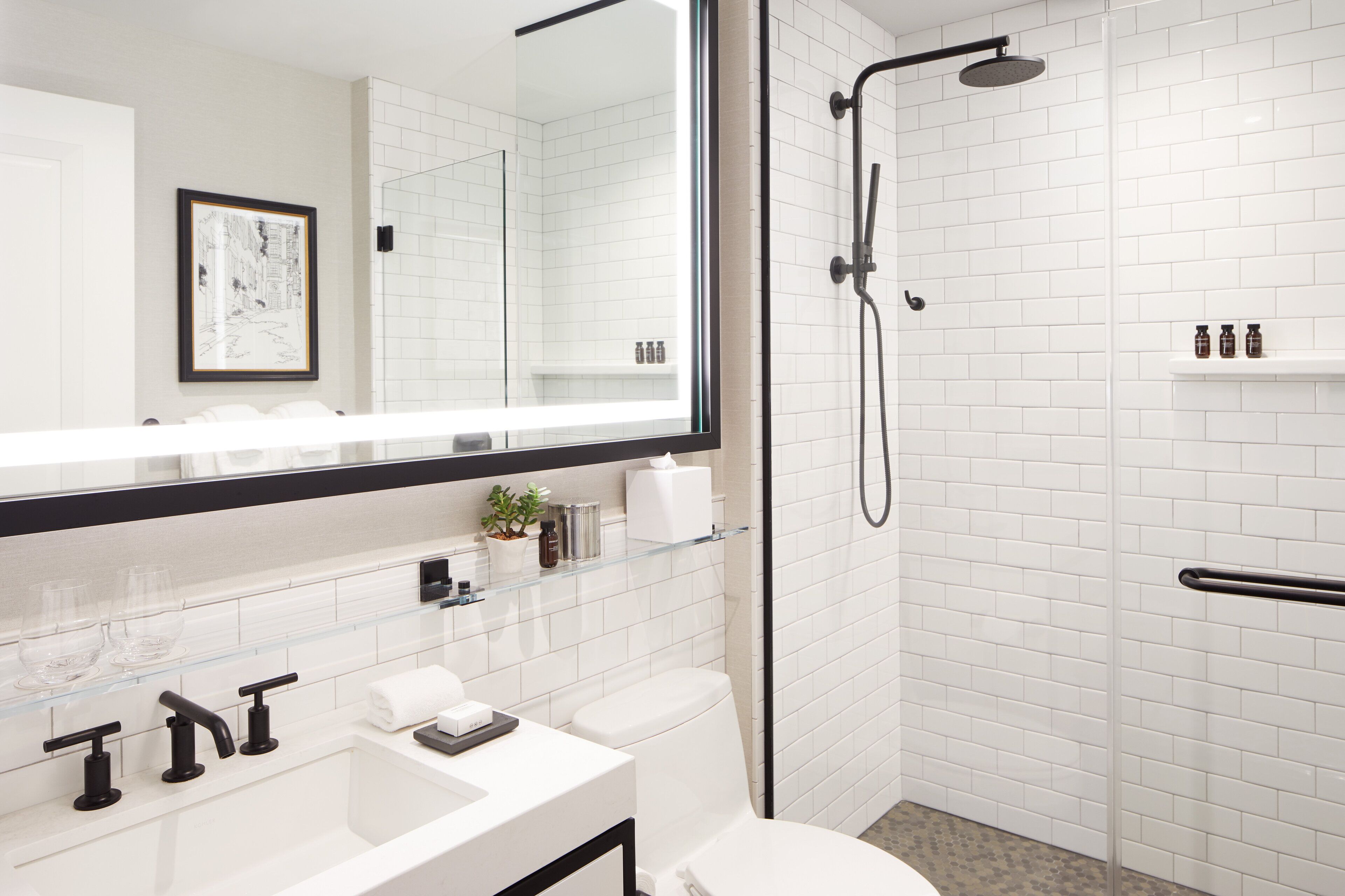 A modern bathroom with a glass shower enclosure, a white sink with black faucets, a large mirror, and a framed picture on the wall.