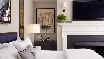 A modern hotel room with a large bed, a flat-screen TV, a fireplace, a nightstand with a lamp, and a framed picture on the wall.