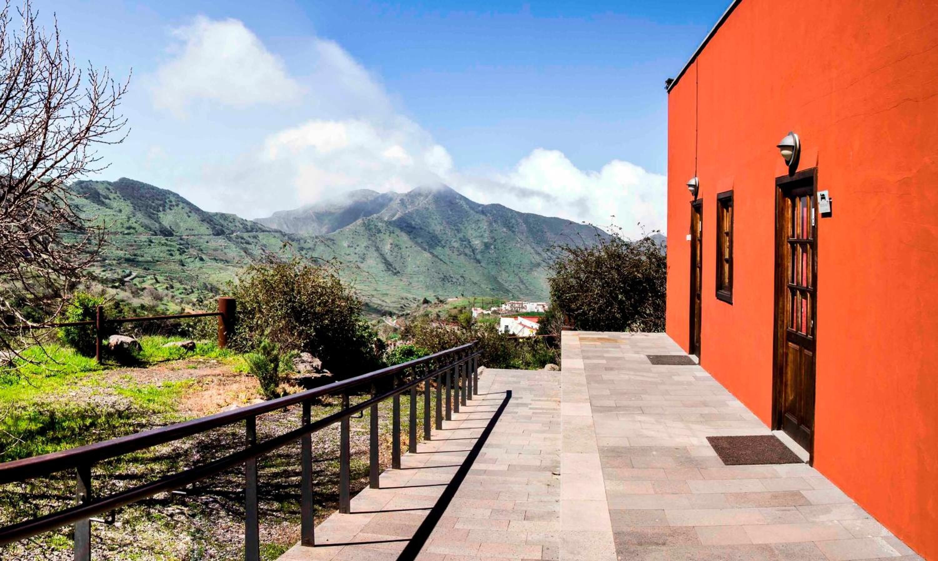 A building with a red exterior, a stone pathway leading to a door, and a view of green hills and mountains in the background.