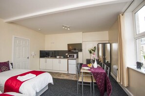 A compact hotel room with a bed, a small kitchenette, a dining table with chairs, and a large window.