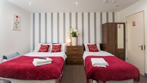 A hotel room with two beds, striped wallpaper, a wooden wardrobe, and a red and white floral arrangement. - Snooze Apartments Flat 52 (Bradford)