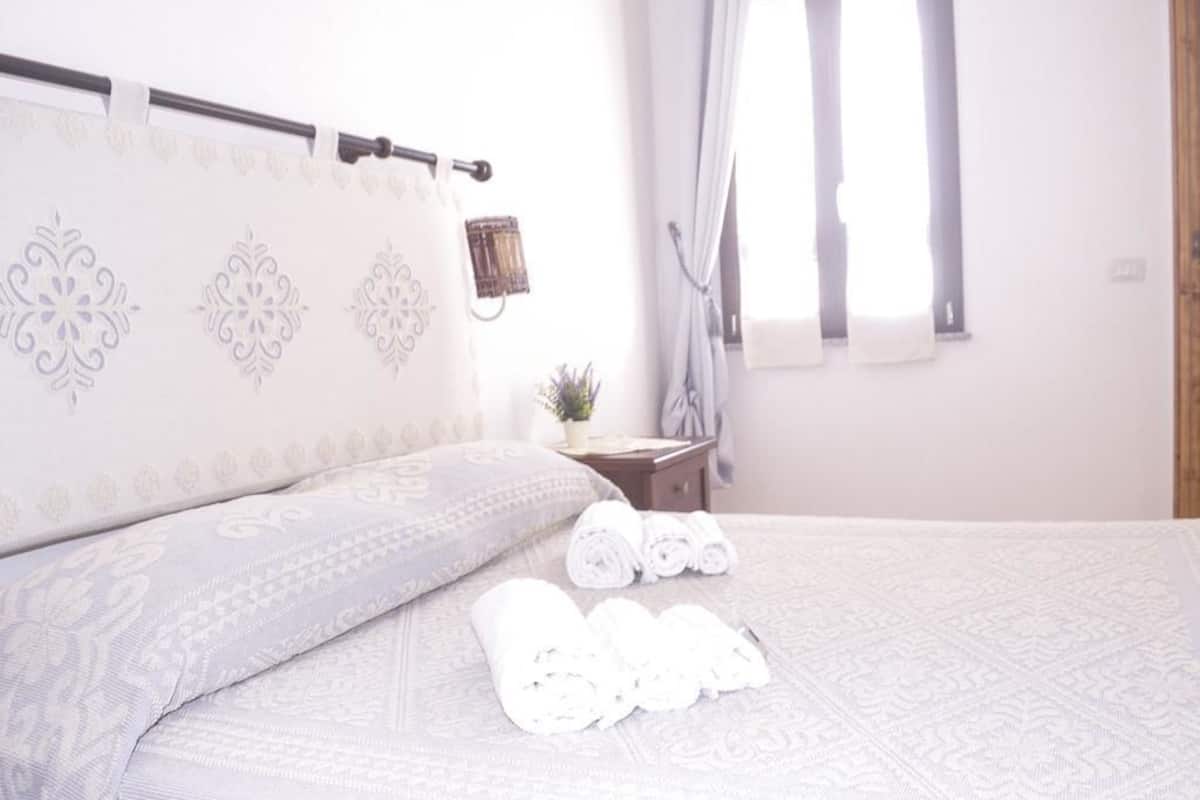 Una cama bien hecha con toallas enrolladas, una mesita de noche con una planta y una ventana con cortinas.