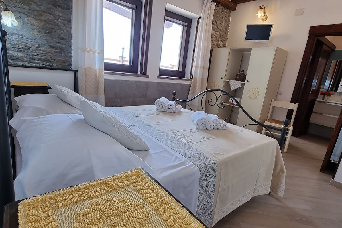 Habitación de hotel con una cama grande, una mesita de noche con un mantel individual amarillo tejido, una pared de piedra y un techo de madera.