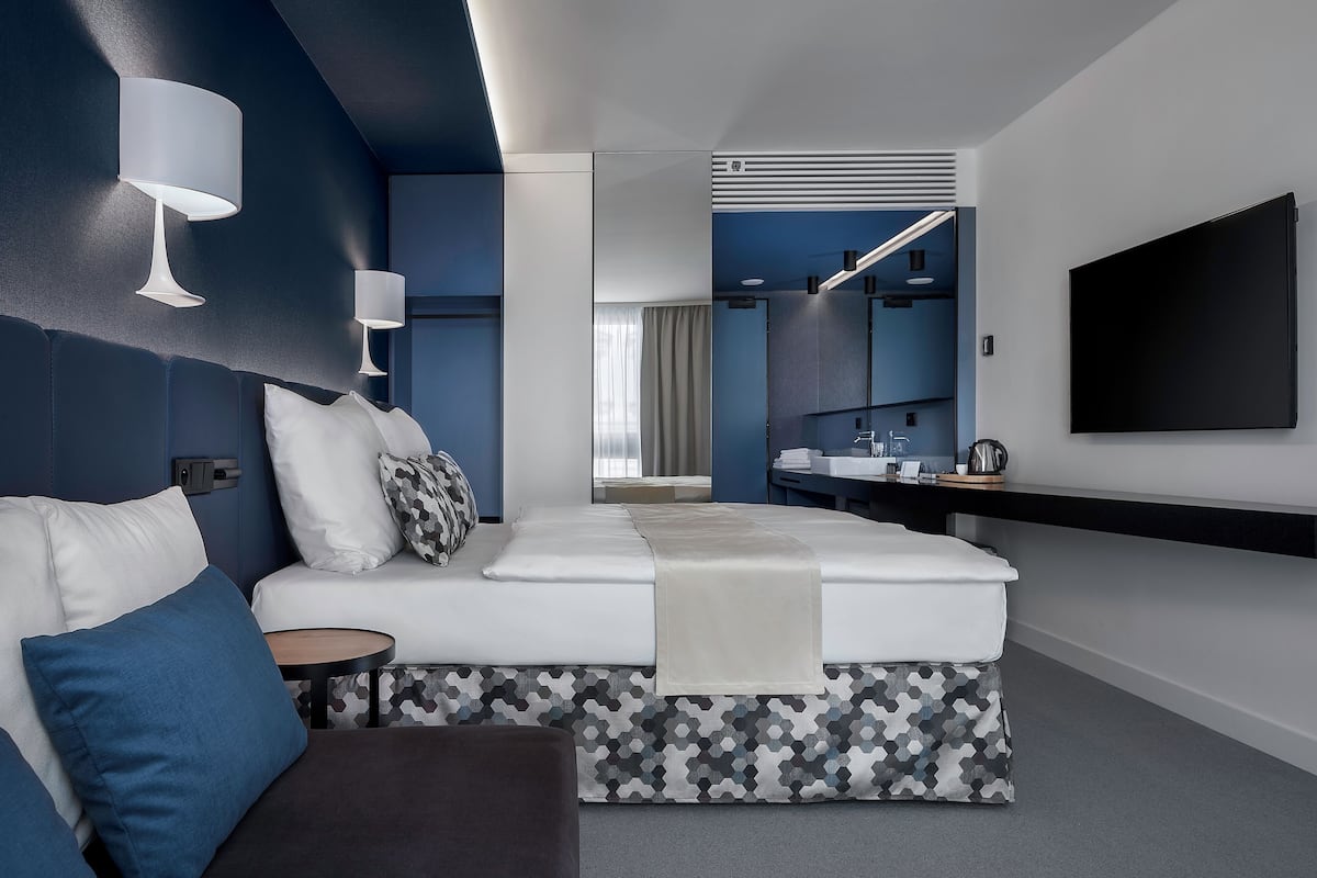 Une chambre d’hôtel moderne, équipée d’un lit, de tables de chevet, d’une télévision et d’une salle de bain visible à travers une porte ouverte.