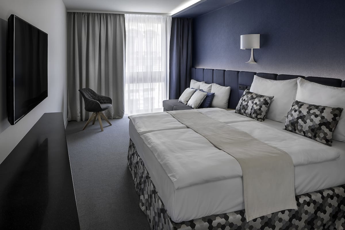 Une chambre d’hôtel moderne dotée d’un grand lit, d’un téléviseur à écran plat, d’un fauteuil et d’une grande fenêtre avec des rideaux.