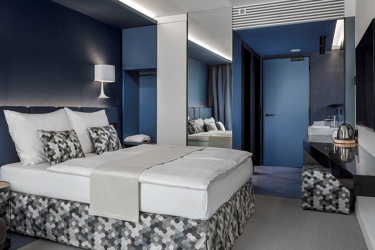Une chambre d’hôtel moderne avec un grand lit, une table de chevet, un miroir et une salle de bain visible à travers une porte ouverte.