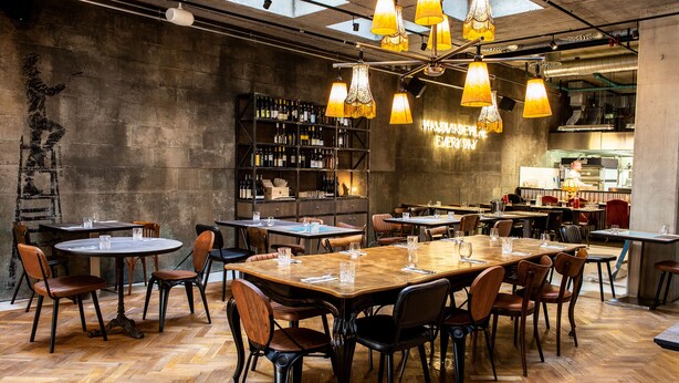 Un restaurant avec des tables et des chaises en bois, un bar avec des étagères remplies de bouteilles et un panneau néon indiquant : « FAIT À LA MAIN TOUS LES JOURS ».