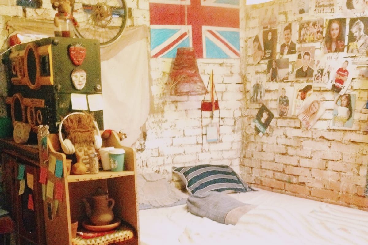 Ein Zimmer mit einem Bett, einem Regal mit diversen Gegenständen, einer Wand mit einer Union Jack Flagge und einer Ziegelwand mit Bildern und Postern.
