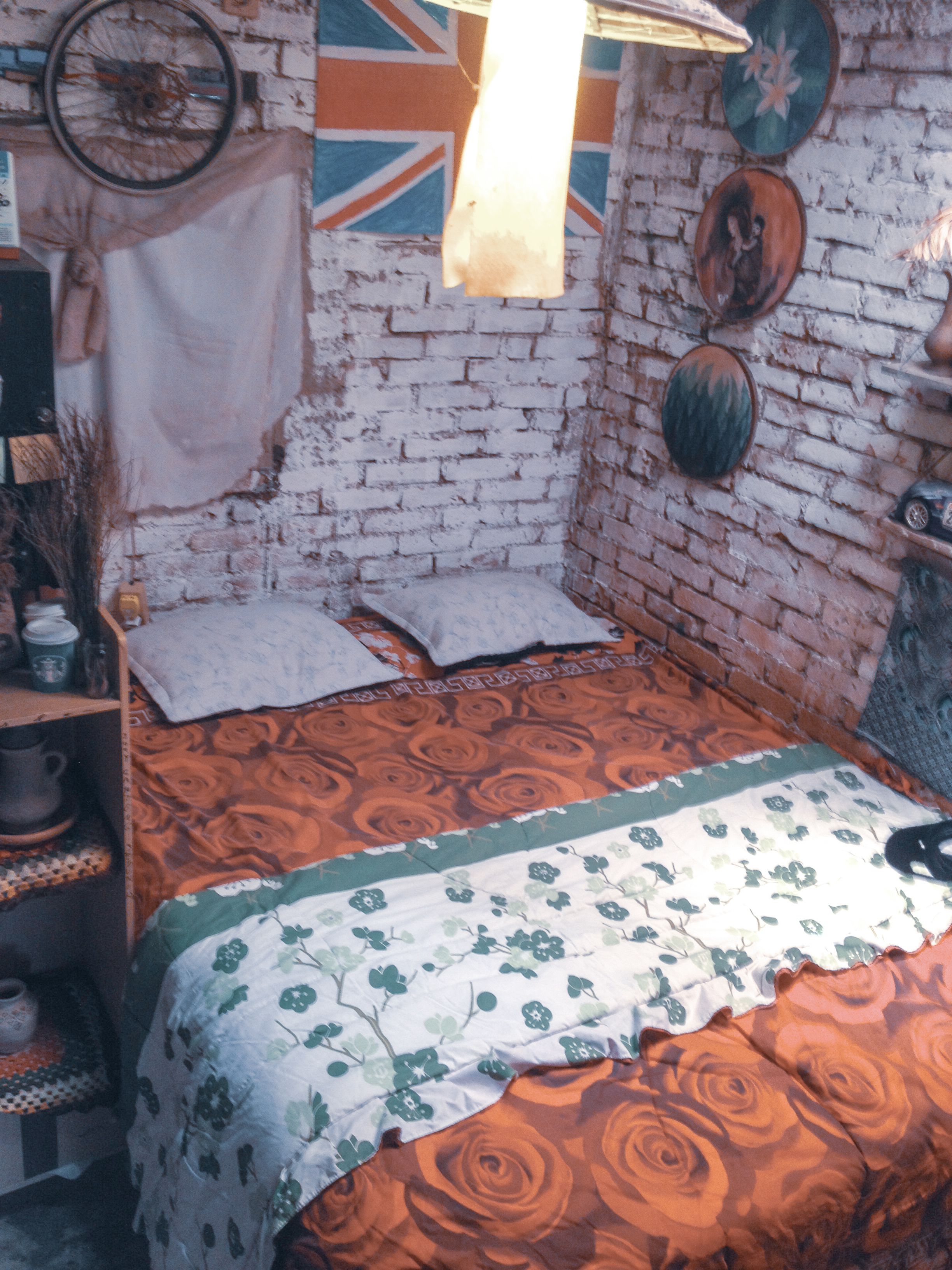 Une pièce avec un mur en briques, un lit recouvert d’une couvre-lit à motifs floraux, un luminaire suspendu et une étagère contenant divers objets.