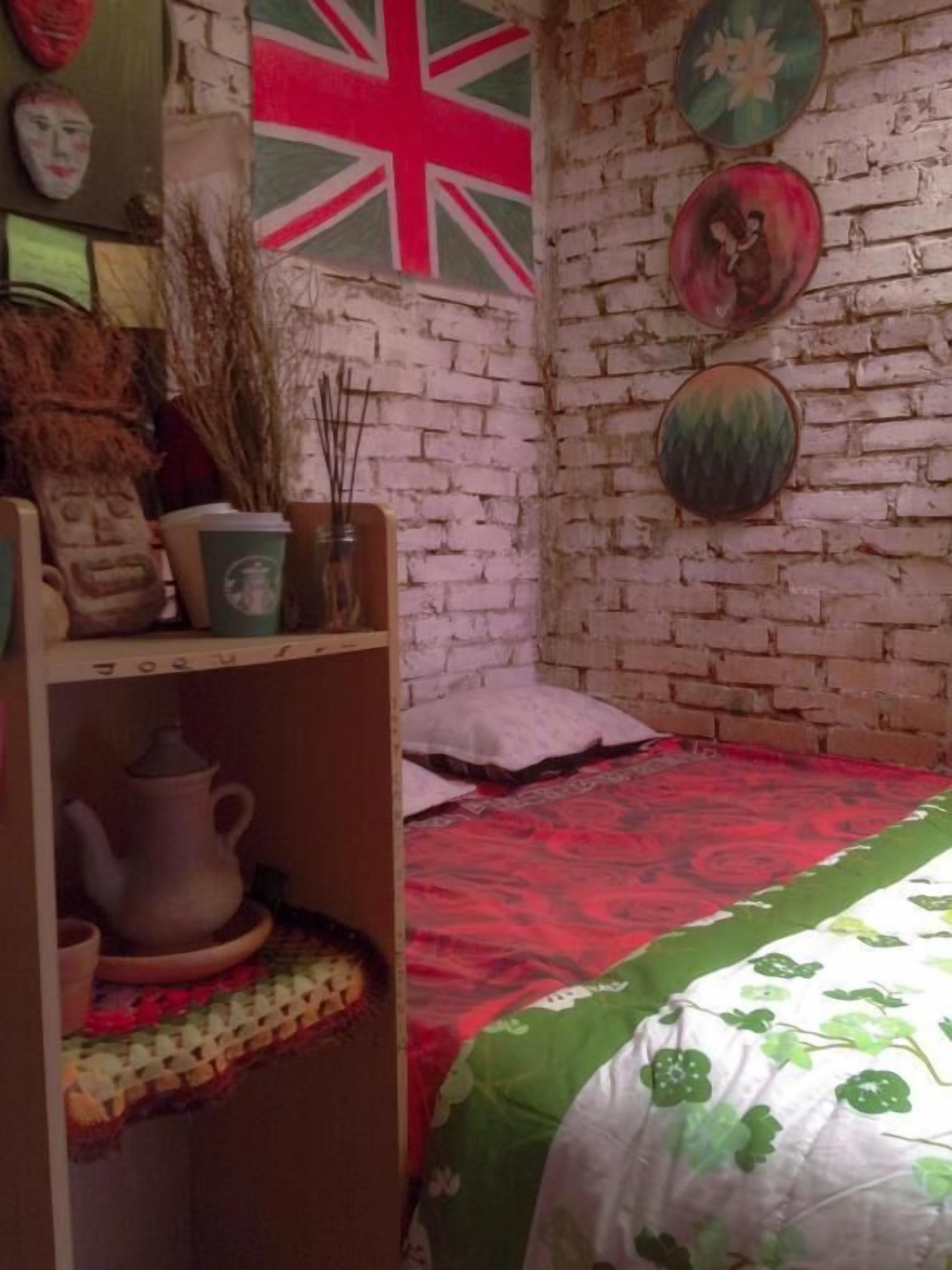 Um quarto com parede de tijolos, uma cama com colcha estampada em vermelho e verde, uma prateleira com uma chaleira e uma bandeira da Union Jack.