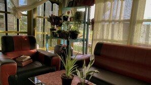 Ein gemütliches Zimmer mit einem rot-schwarzen Sofa, einem Glastisch mit Topfpflanzen und einem Fenster mit Spitzenvorhängen.