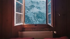 A cozy cabin room with a wooden ceiling, a window with a view of snow-covered mountains, a bed with red bedding, and a small bedside table. - Mountain Hostel Gimmelwald (Lauterbrunnen)