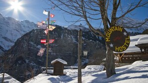 A mountain hostel sign with multiple flags, a wooden structure, and snow-covered mountains in the background. - Mountain Hostel Gimmelwald (Lauterbrunnen)