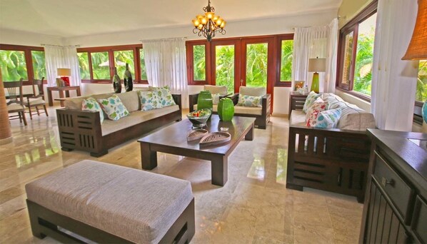 A spacious living room with a sofa set, a coffee table, and a dining table. Large windows with curtains provide natural light and a view of greenery outside.