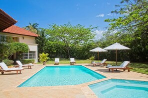 A swimming pool with lounge chairs and umbrellas, surrounded by greenery and a building with a red-tiled roof.