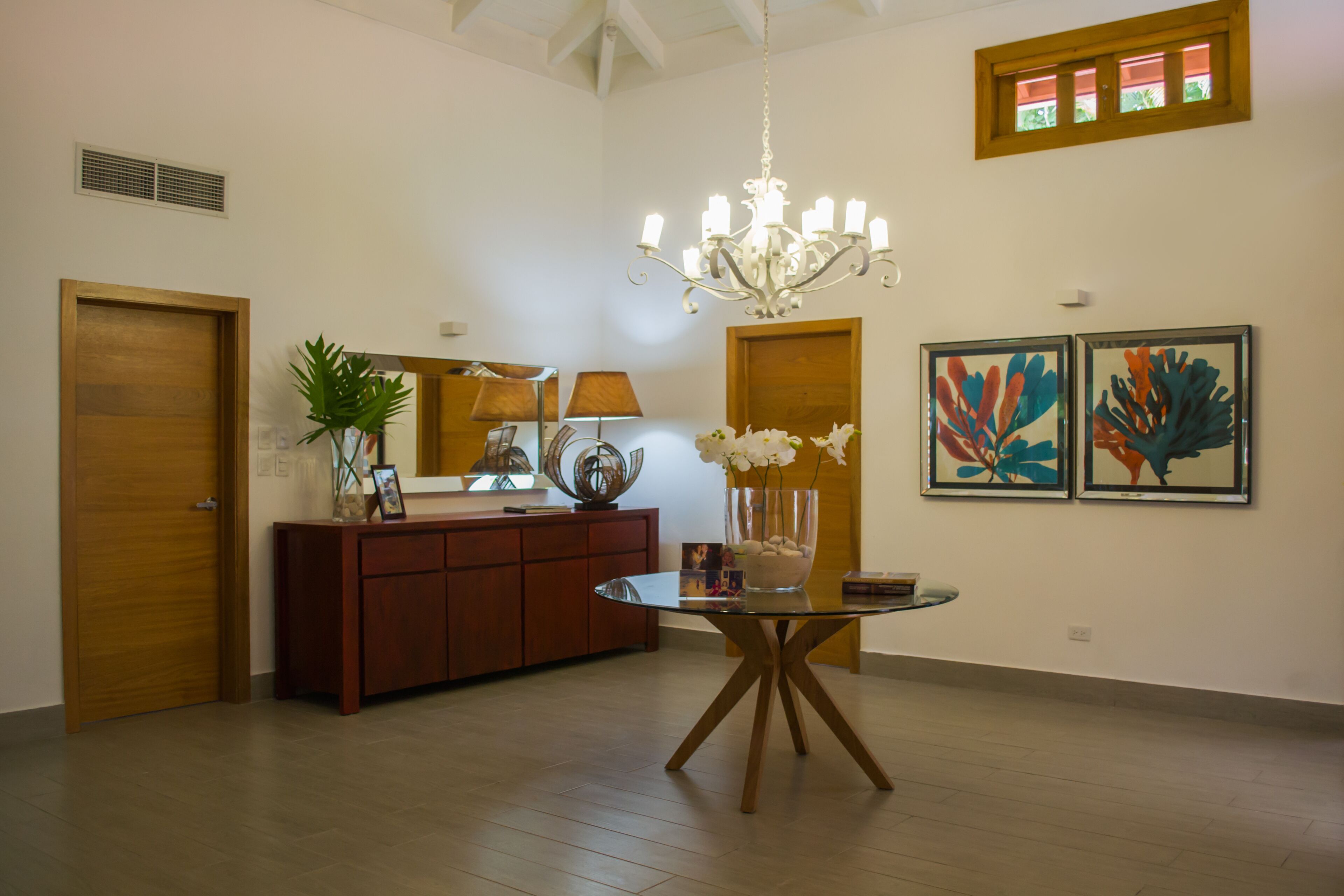 A spacious room with a wooden floor, a glass-top dining table, a wooden cabinet, a chandelier, and two framed artworks on the wall.