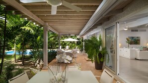 Un patio techado al aire libre con piscina, mobiliario blanco y abundante vegetación.