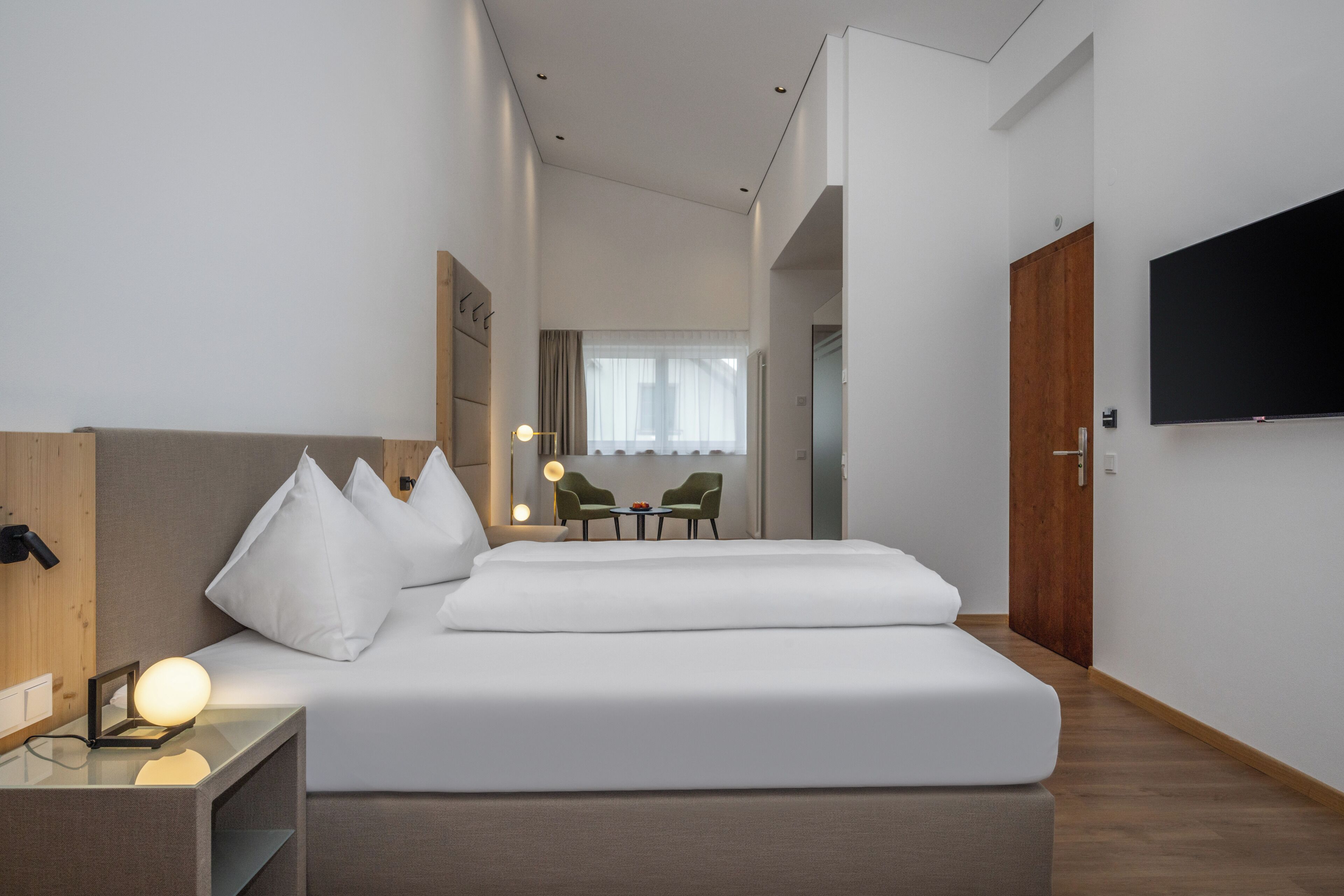 Una habitación de hotel moderna con una cama grande, mesita de noche, lámpara y televisor montado en la pared.