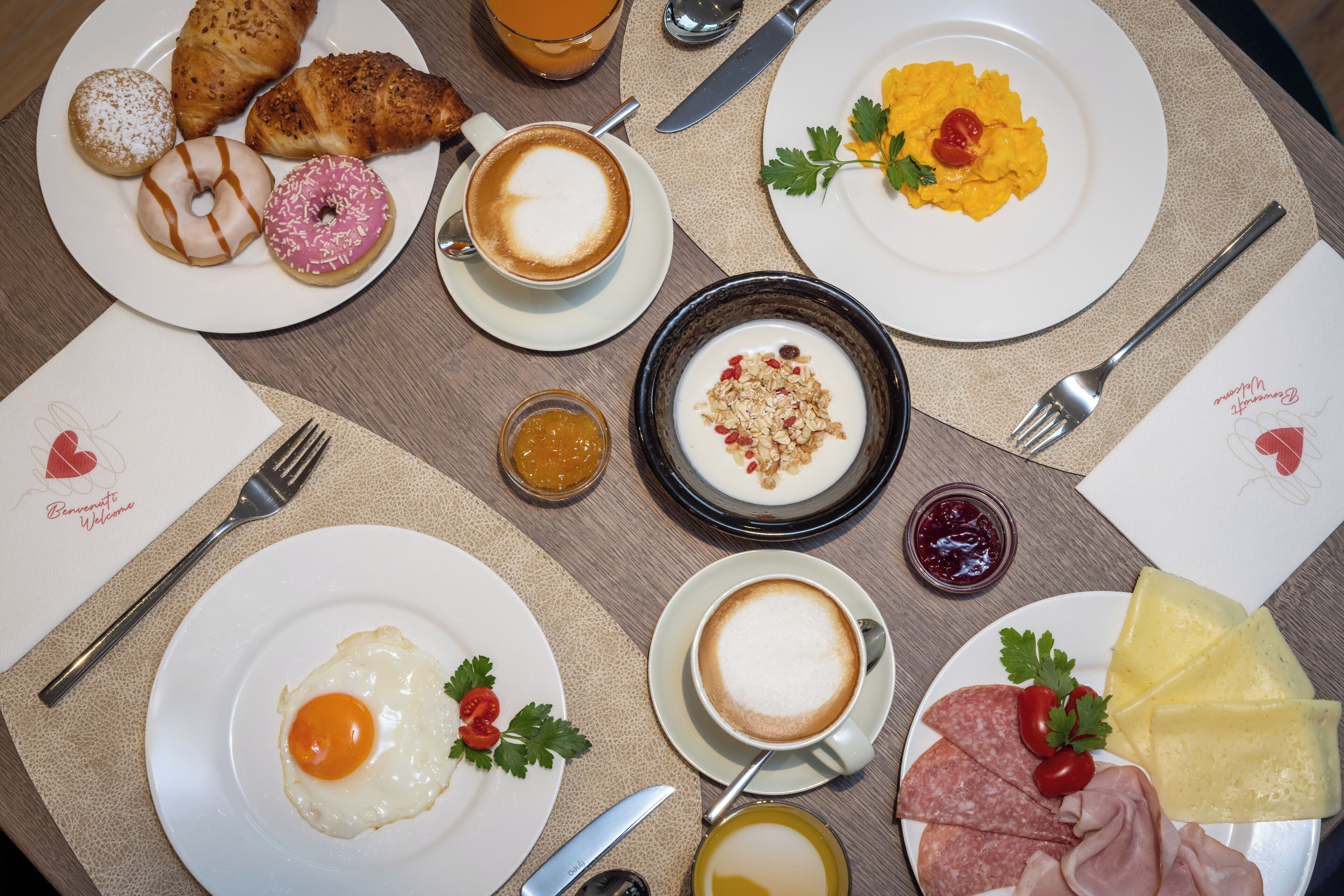 Un desayuno con cruasanes, donas, café, huevos revueltos, yogur, queso y diferentes untables en platos.