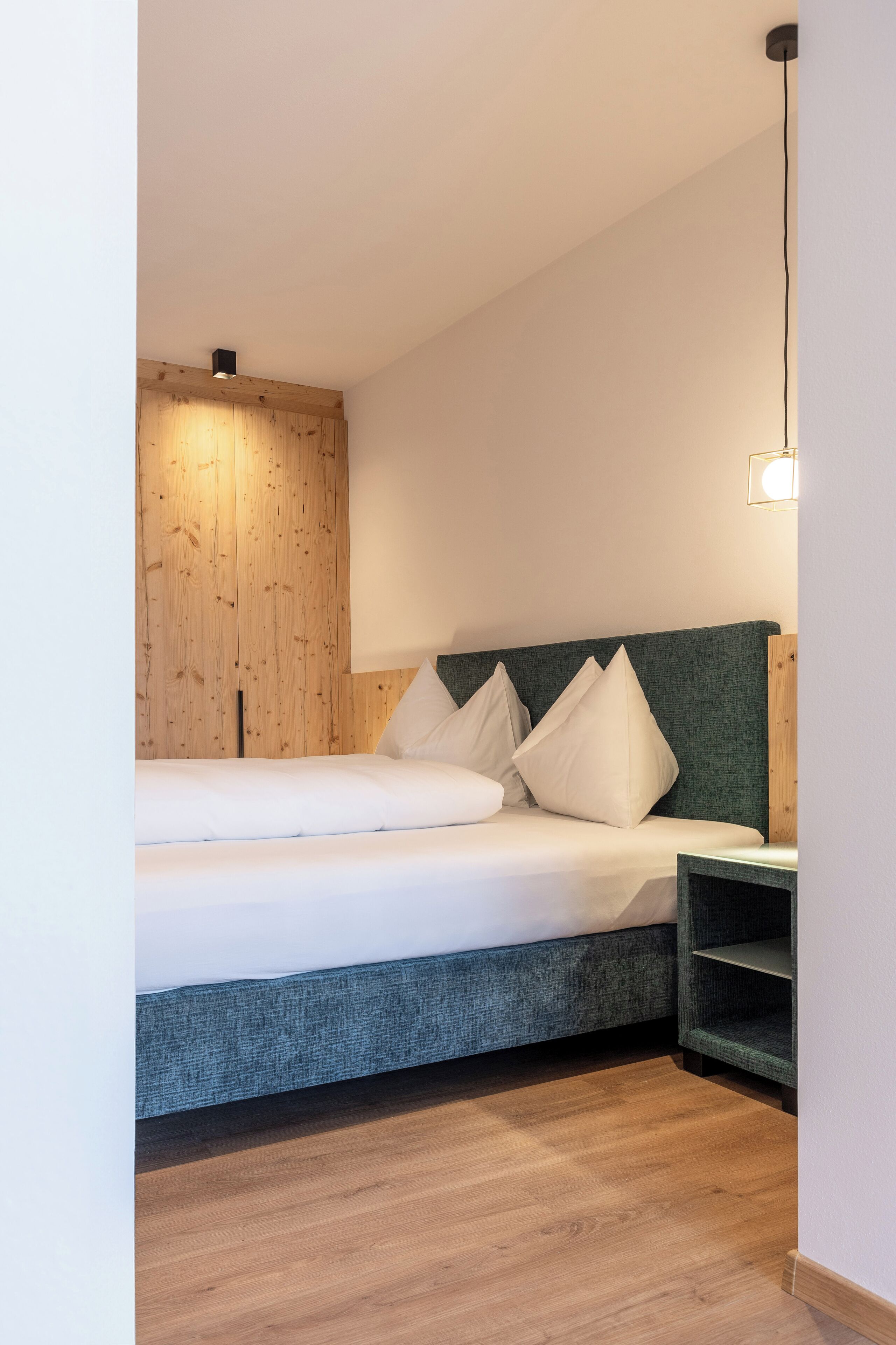 Un dormitorio moderno con pared de madera, cabecera tapizada en verde, cama blanca con almohadas y mesita de noche.