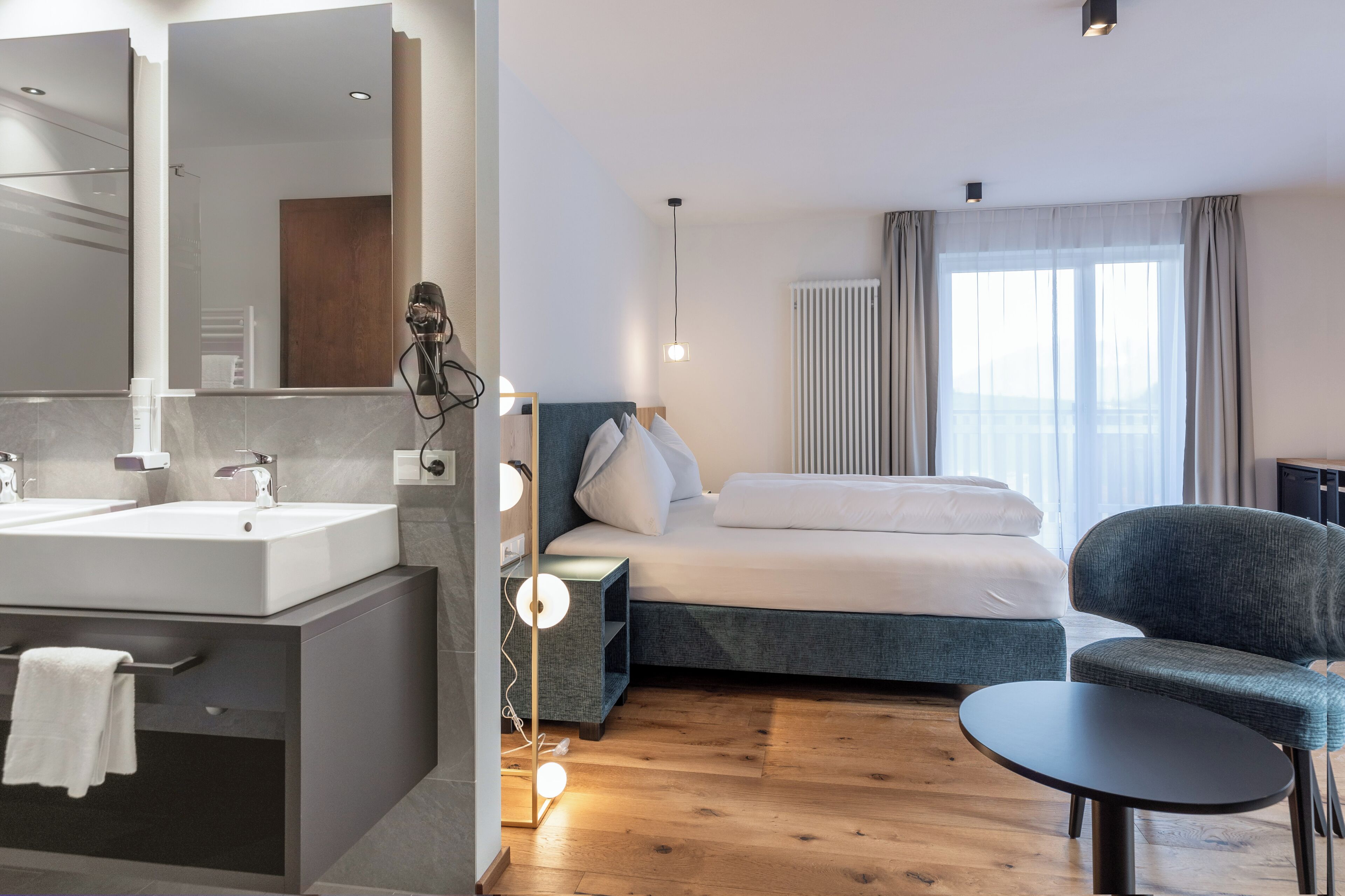 Una habitación de hotel moderna con una cama grande, una mesita de noche, una silla, una pequeña mesa, un baño con lavabo y espejo, y una ventana con cortinas.