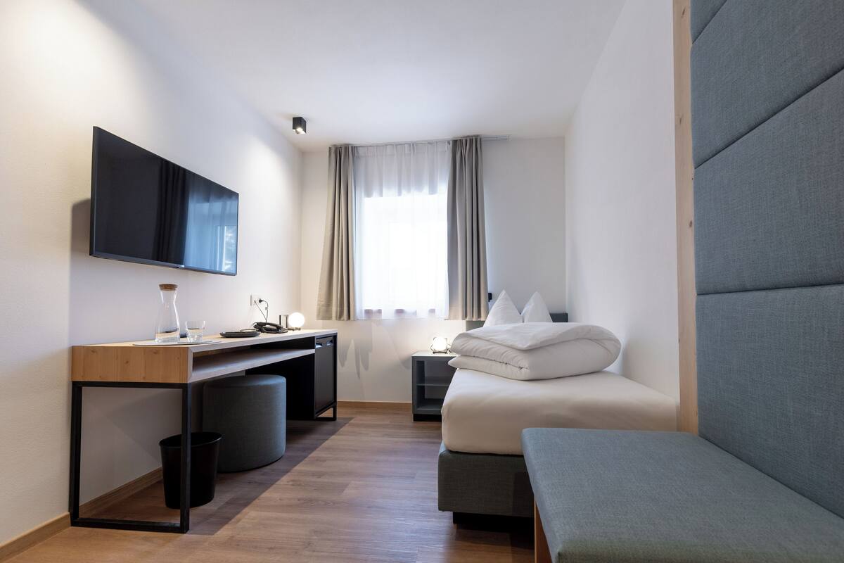 Ein modernes Hotelzimmer mit Wand-Fernseher, einem Holztisch mit Stuhl, einem Bett mit grauem gepolsterten Kopfteil und einem Nachttisch mit Lampe.