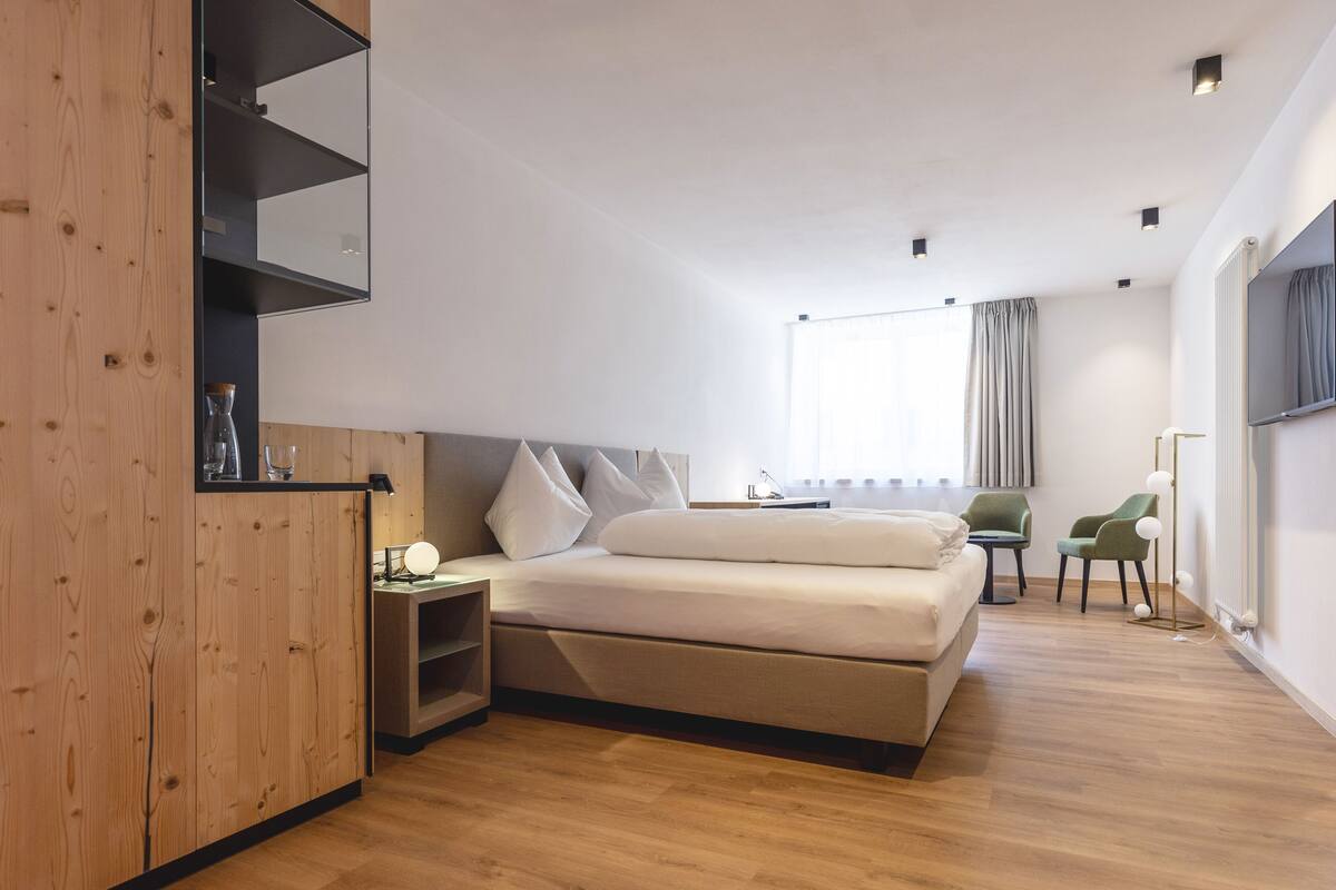 Ein modernes Hotelzimmer mit einem Holzkopfteil, einem großen Bett, einem Nachttisch, einem Fernseher und einem Fenster mit Vorhängen.