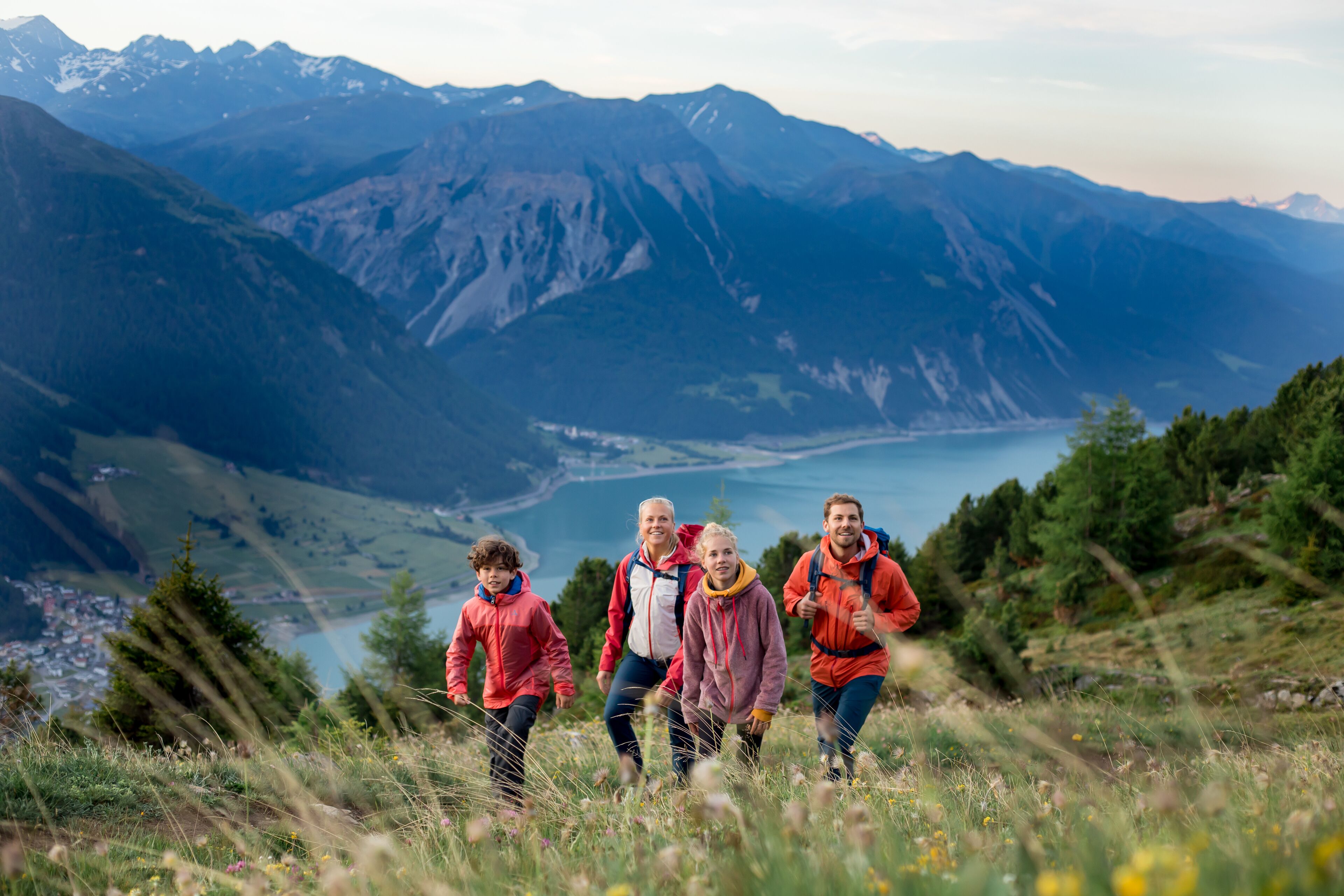 A group of people hiking on a mountain trail with a lake and mountains in the background.