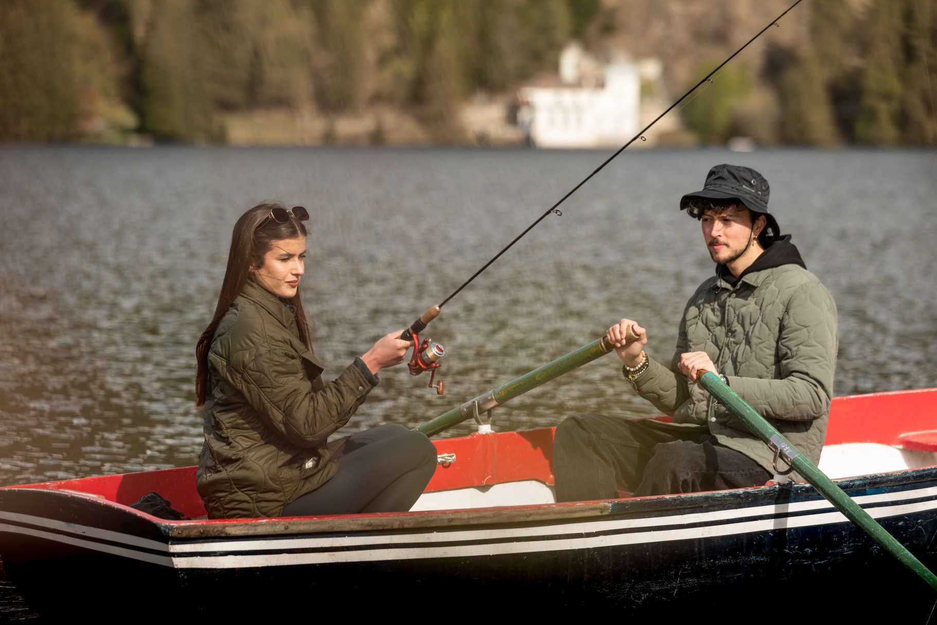 Two people fishing in a small boat on a lake with a house in the background.