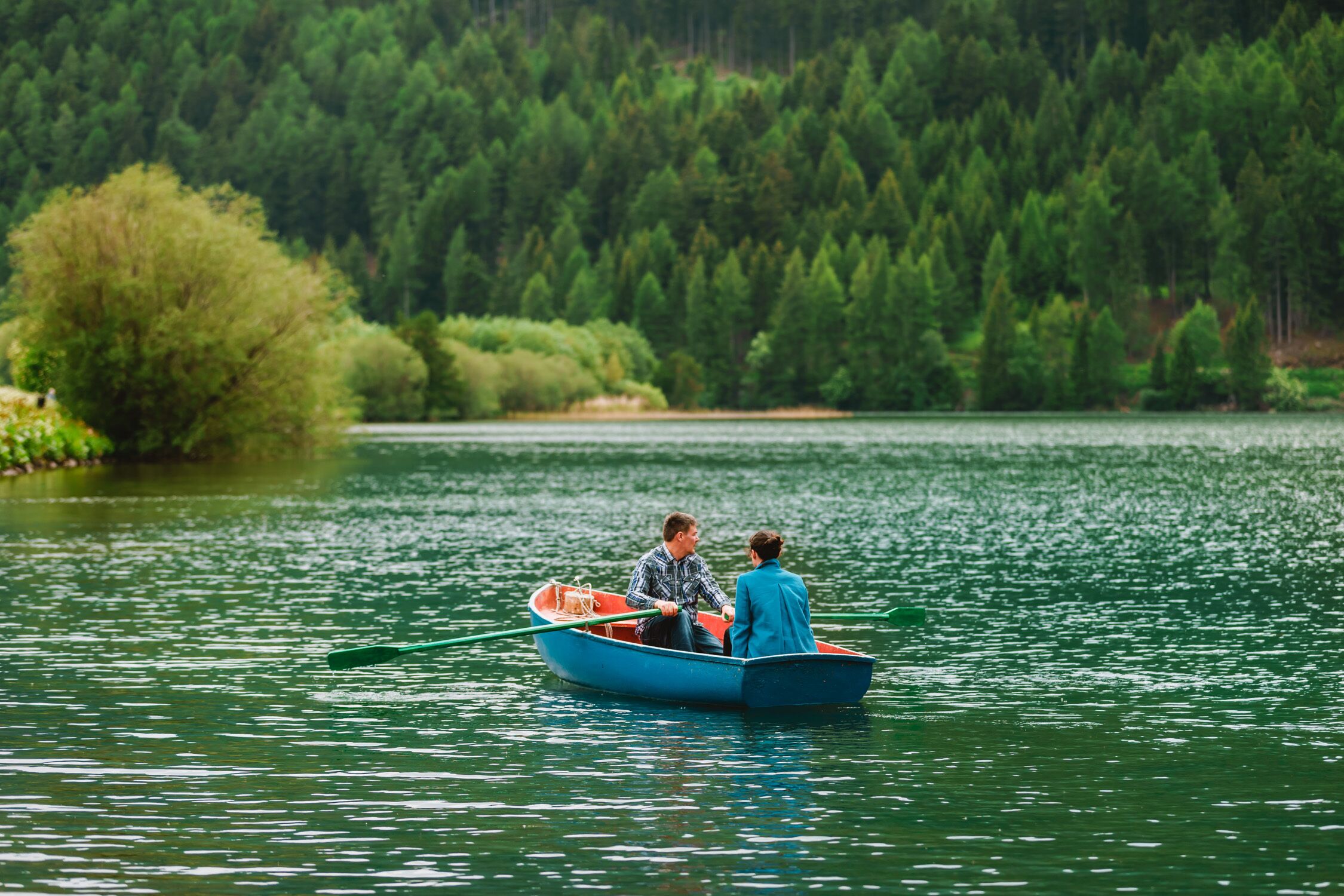 Two people in a small boat on a calm lake surrounded by dense forest.