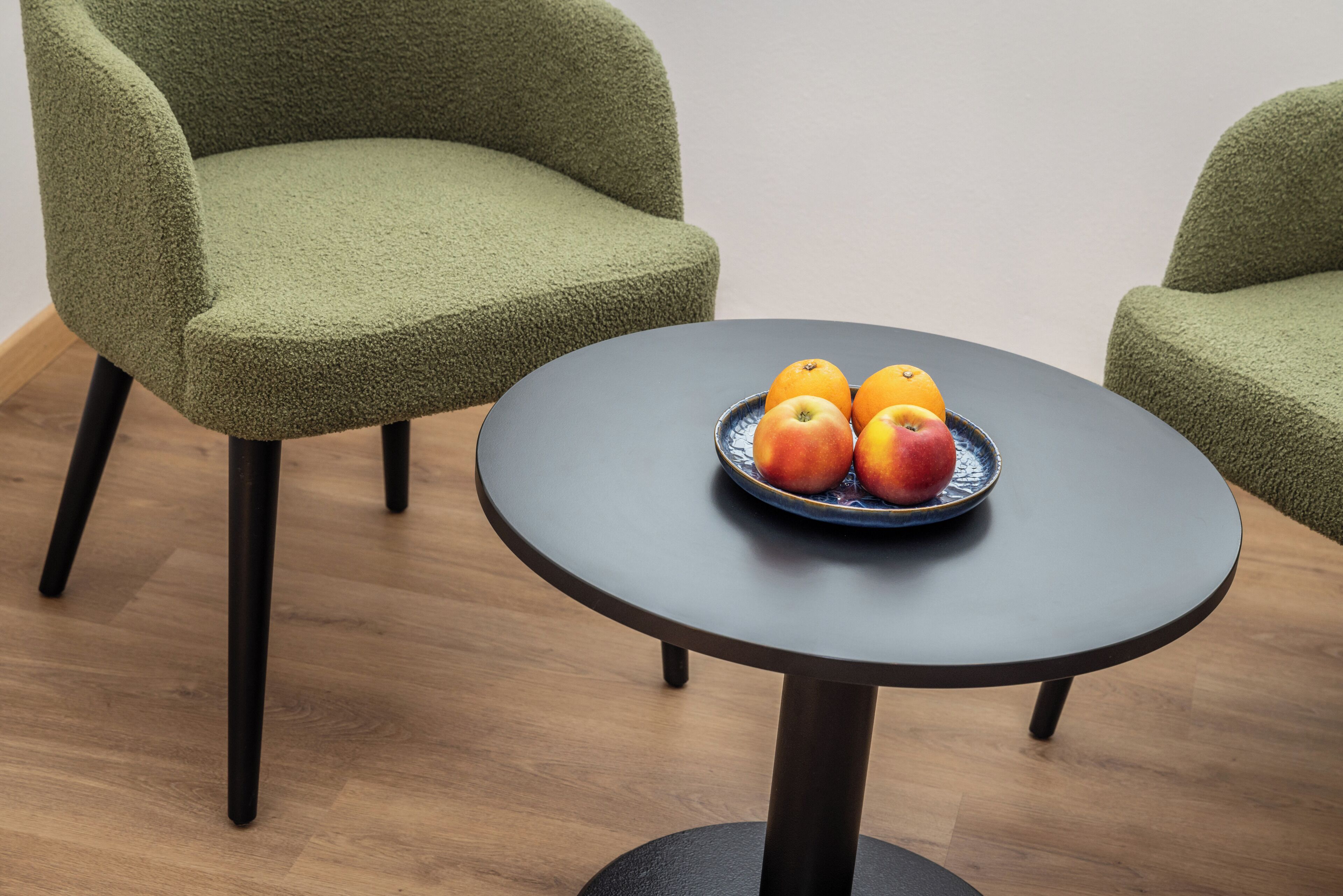 A round table with a dark surface and a blue plate holding three apples and two oranges.