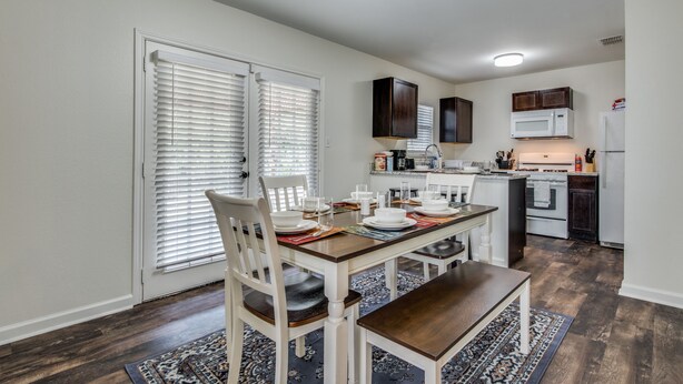 A dining area with a wooden table set for four, white chairs, and a patterned rug.