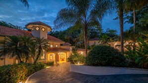 A well-lit, spacious entrance with a curved driveway leading to a building with a tiled roof and a central tower. - Harmon House (Jacó)