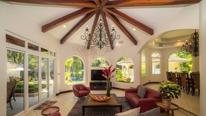 A spacious living room with a red sofa, a wooden coffee table, and a chandelier. There are large windows offering views of a garden and a pool. - Harmon House (Jacó)