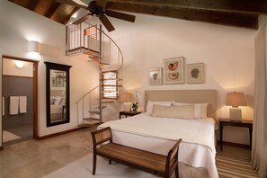 Ein Schlafzimmer mit einem großen Bett, einer hölzernen Bank, einer Wendeltreppe, einem Deckenventilator und gerahmten Bildern an der Wand.